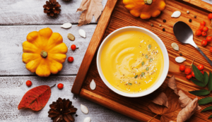 pomarańczowa zupa krem w białej misce stojącej na drewnianej desce wśród jesiennych dekoracji - tytułowa.